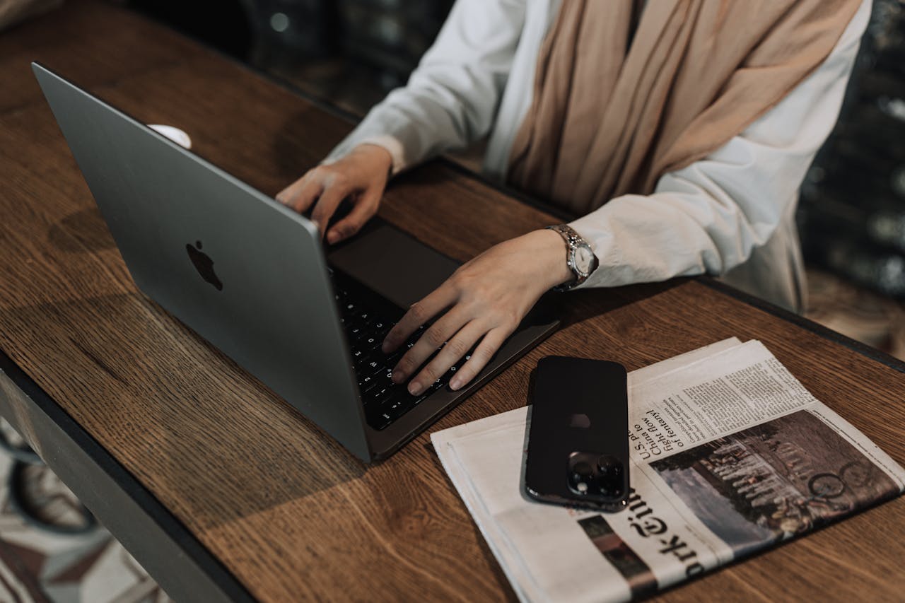 Woman typing on a laptop, with a smartphone and newspaper nearby, in a cozy indoor setting.