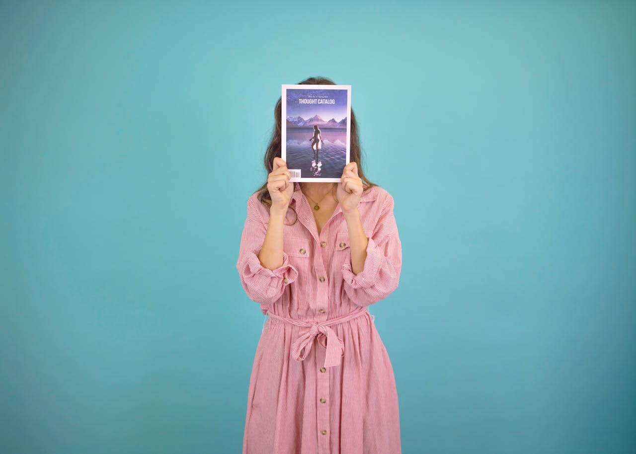 A woman in a pink dress holds a magazine in front of her face against a blue background.
