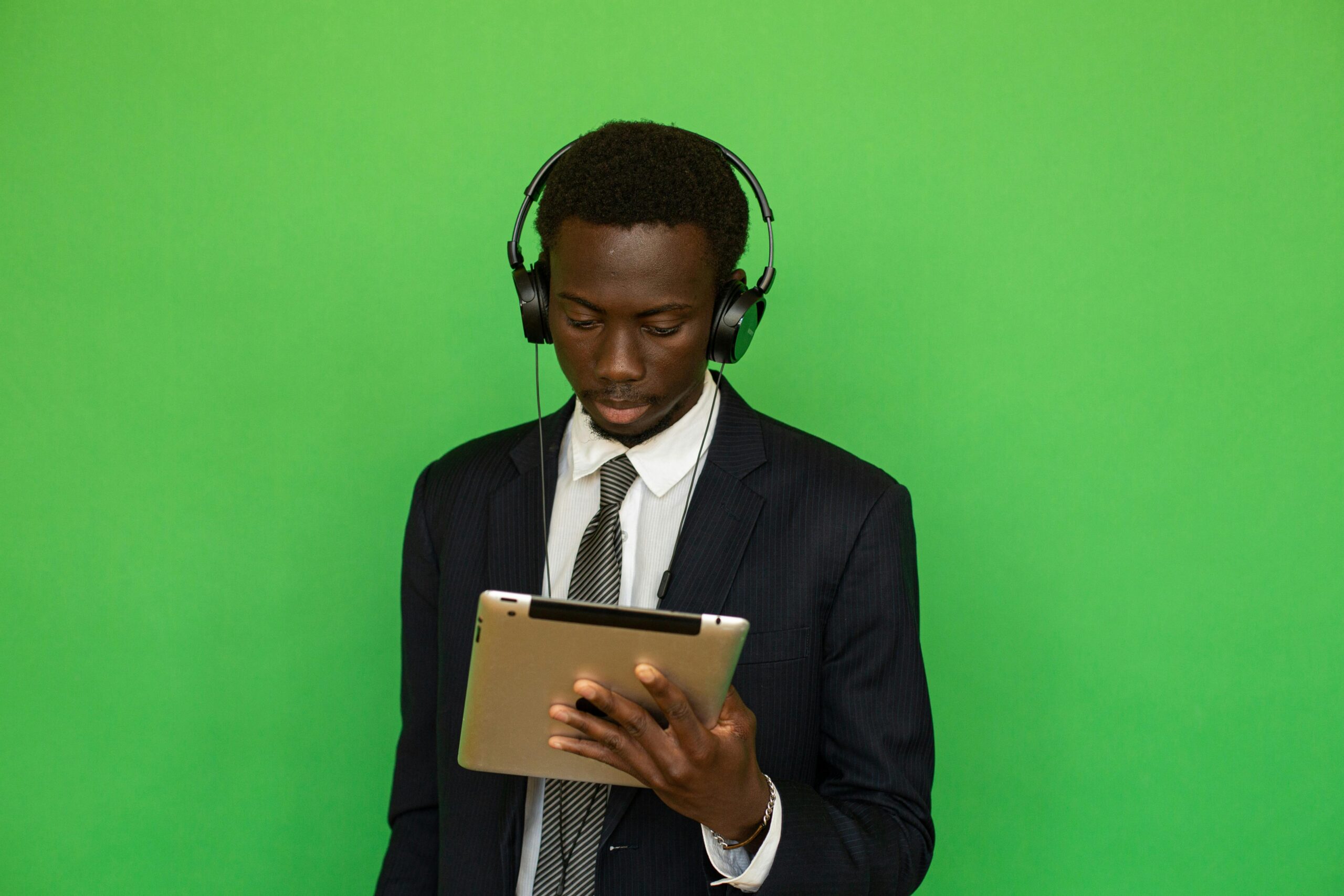 Focused businessman in suit using a tablet with headphones, isolated on a vibrant green background.
